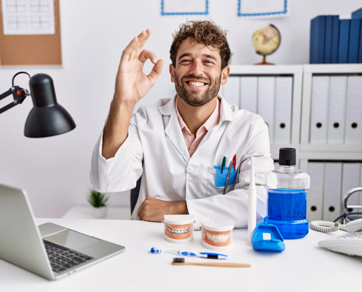 Smiling male dentist in white coat making an OK hand gesture at desk with dental models and oral care tools.