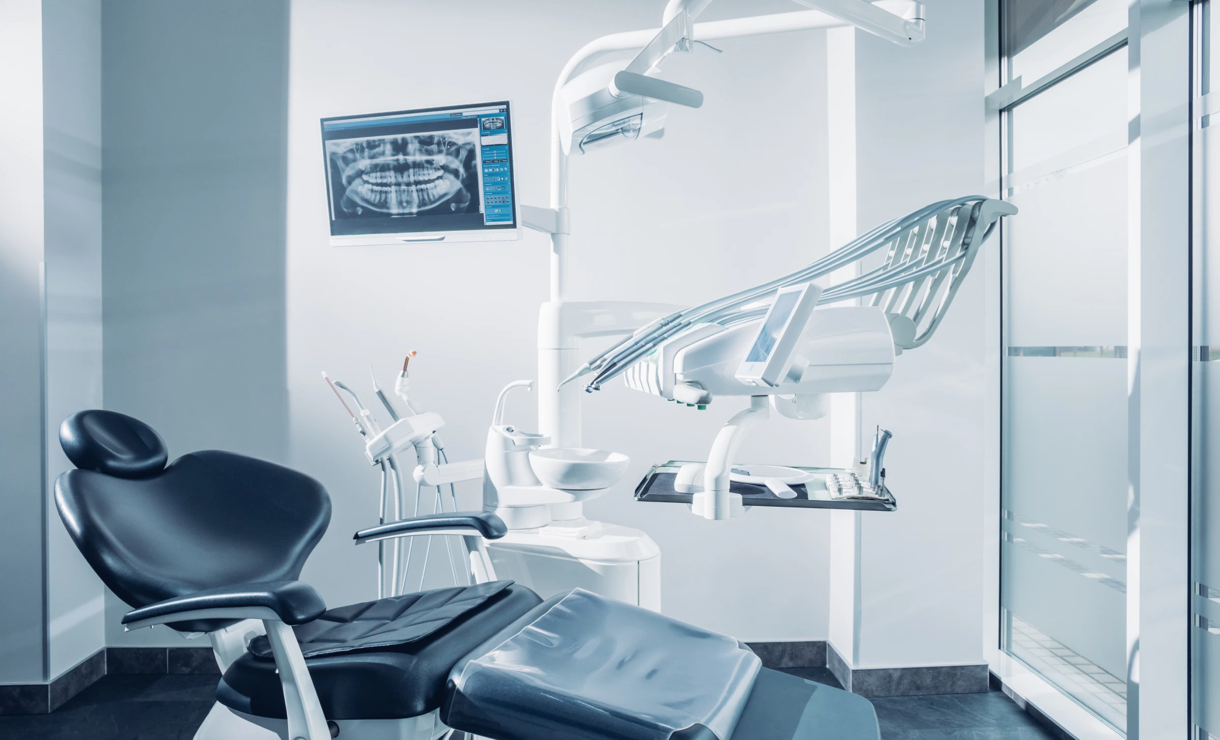 Modern dental examination room with black dental chair, overhead light, dental instruments, and a monitor displaying a dental X-ray.