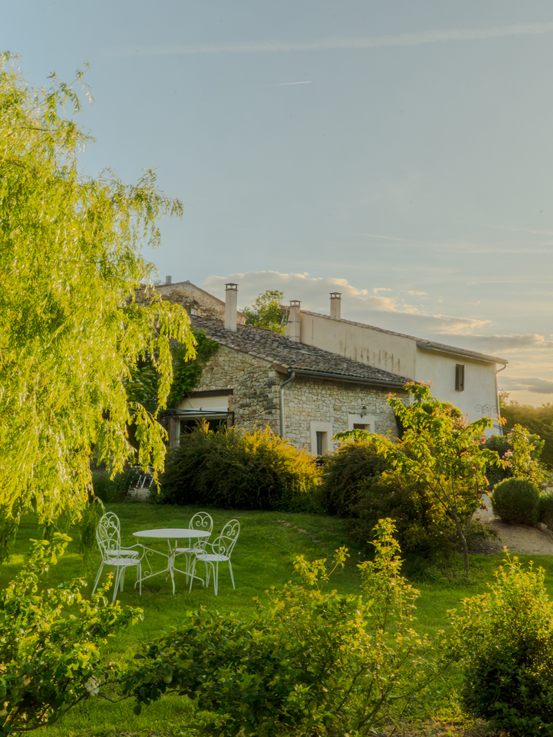 Stone house with wooden door surrounded by lush garden and blooming pink roses under blue sky.