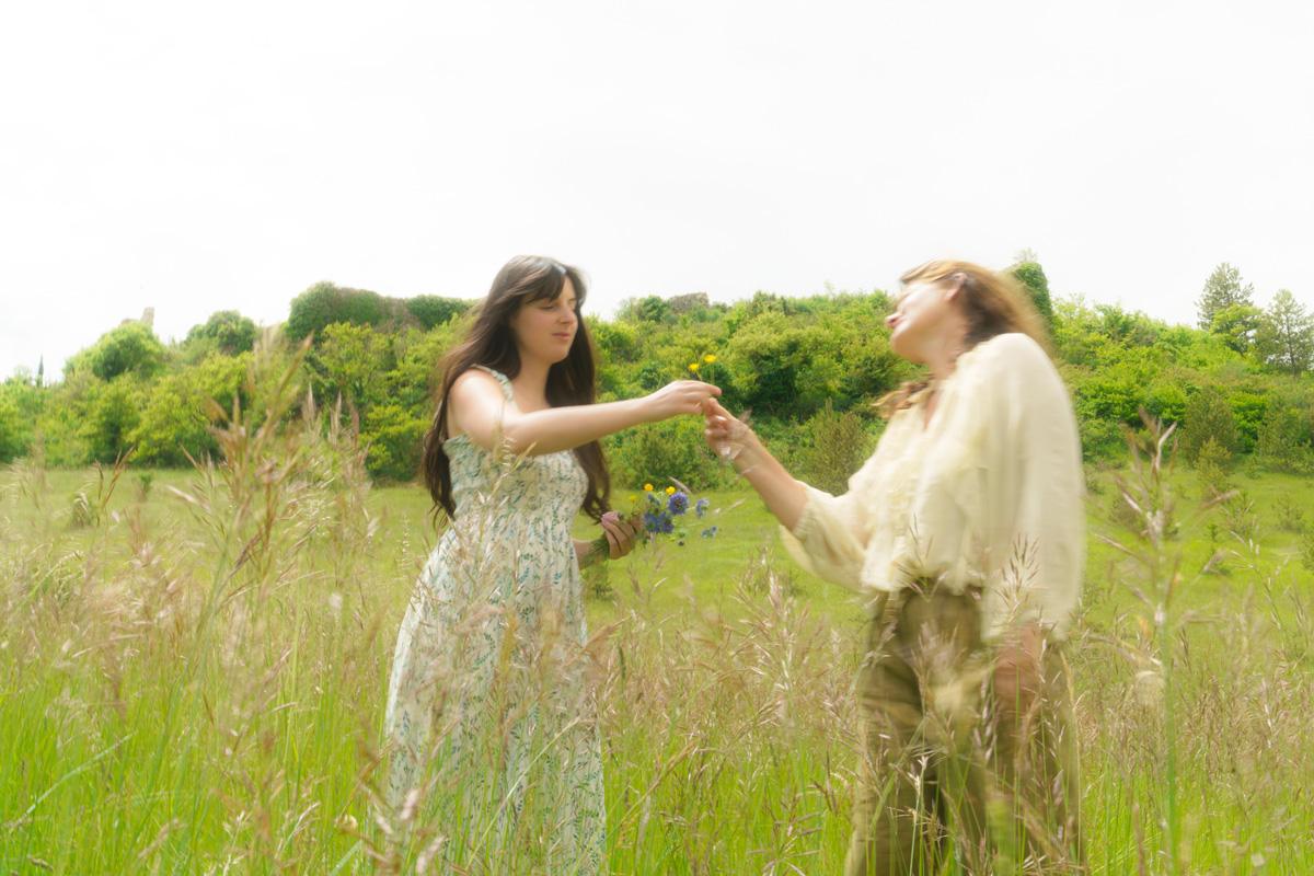 Two women in a grassy field exchanging small flowers on a bright, cloudy day.