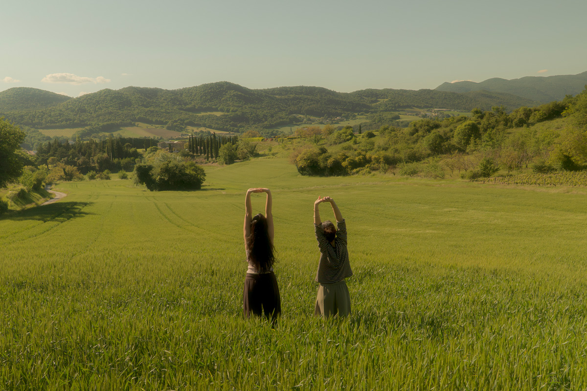 Two people standing in a green field stretching their arms upward with hills in the background.