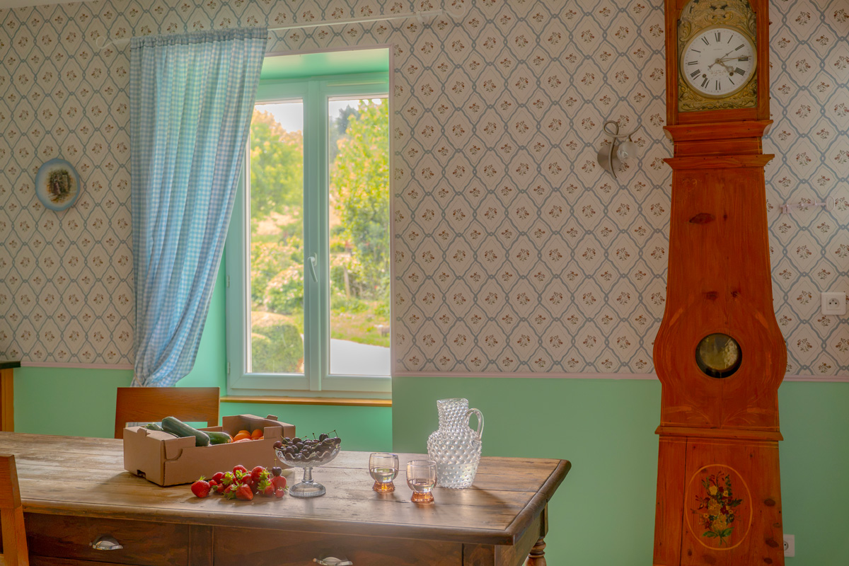 Wooden table with fruit and a glass pitcher in a bright room with patterned wallpaper and a tall clock.