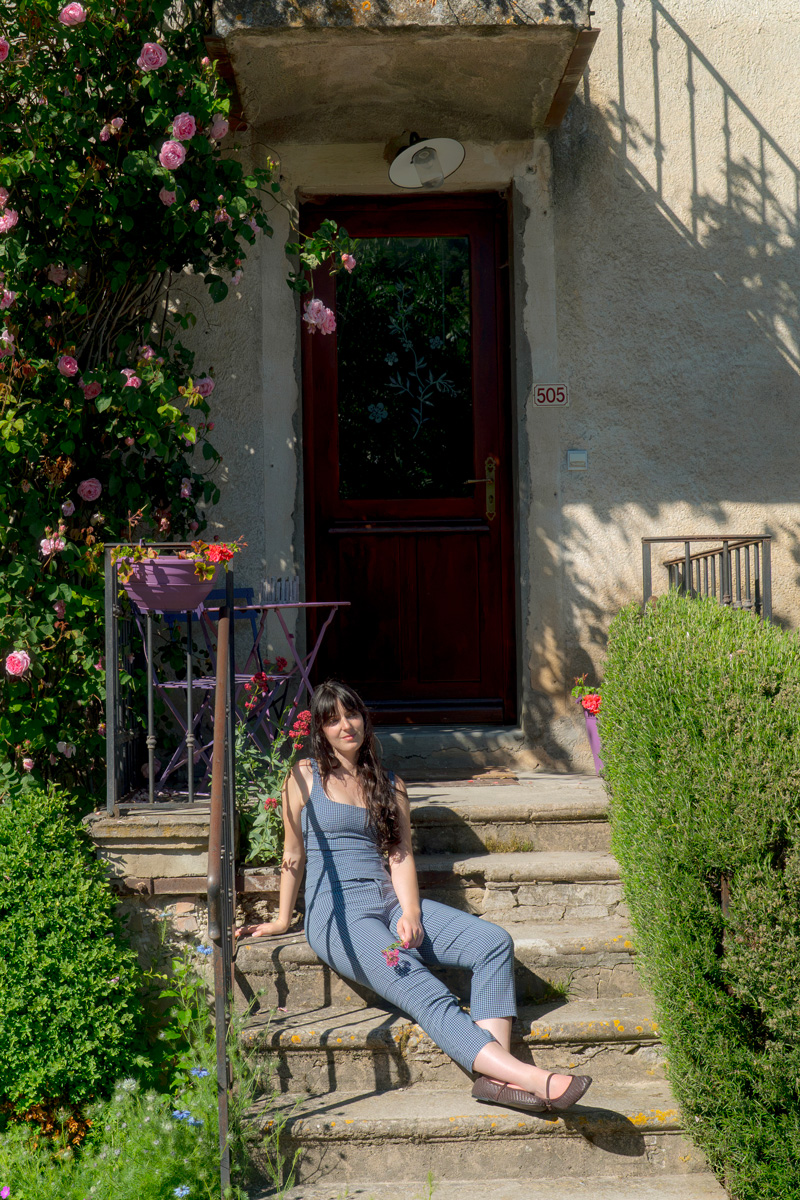 Woman in blue checkered outfit sitting on outdoor steps near pink roses and a wooden door.