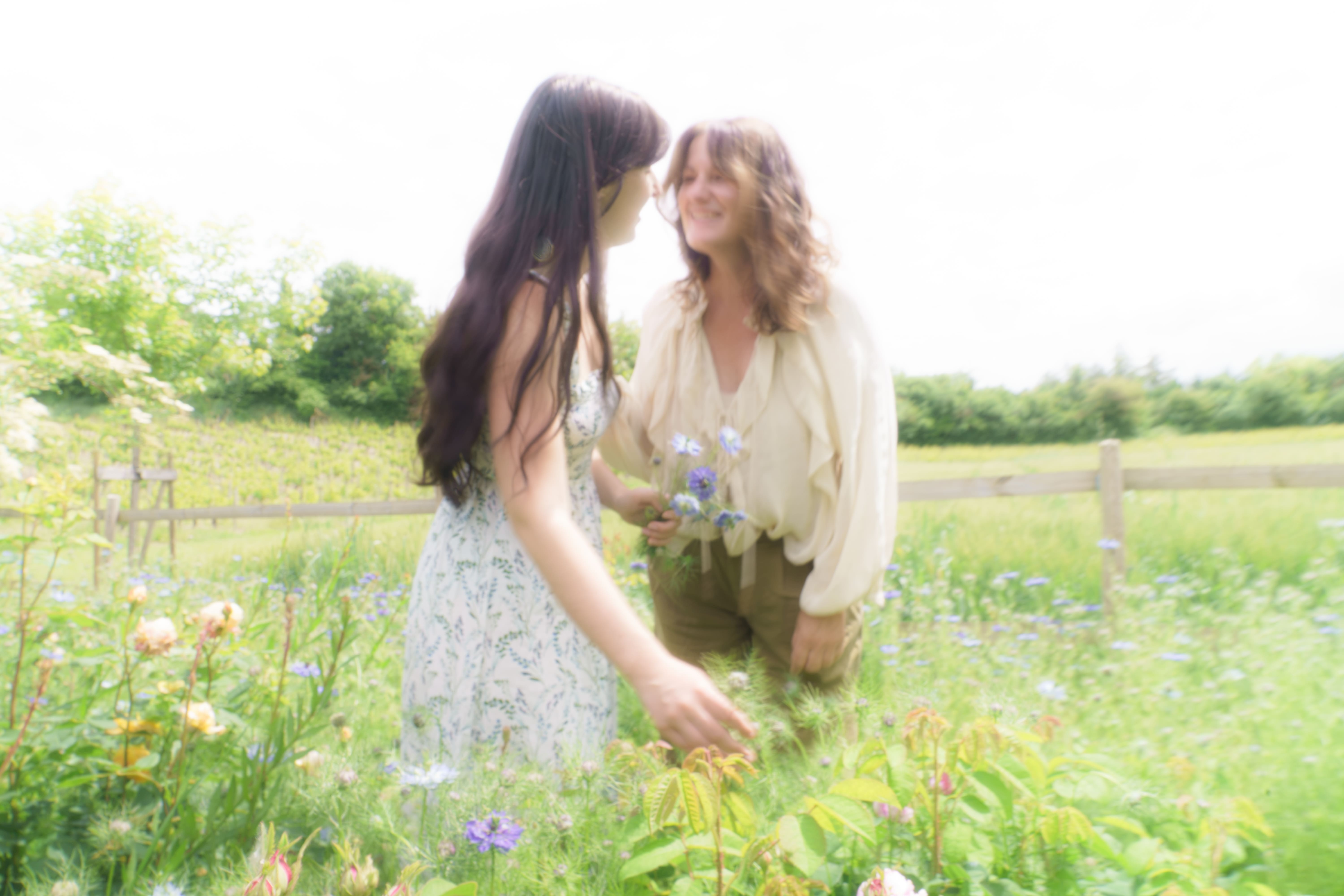 Two women smiling and holding flowers in a sunlit garden with greenery and wooden fence.