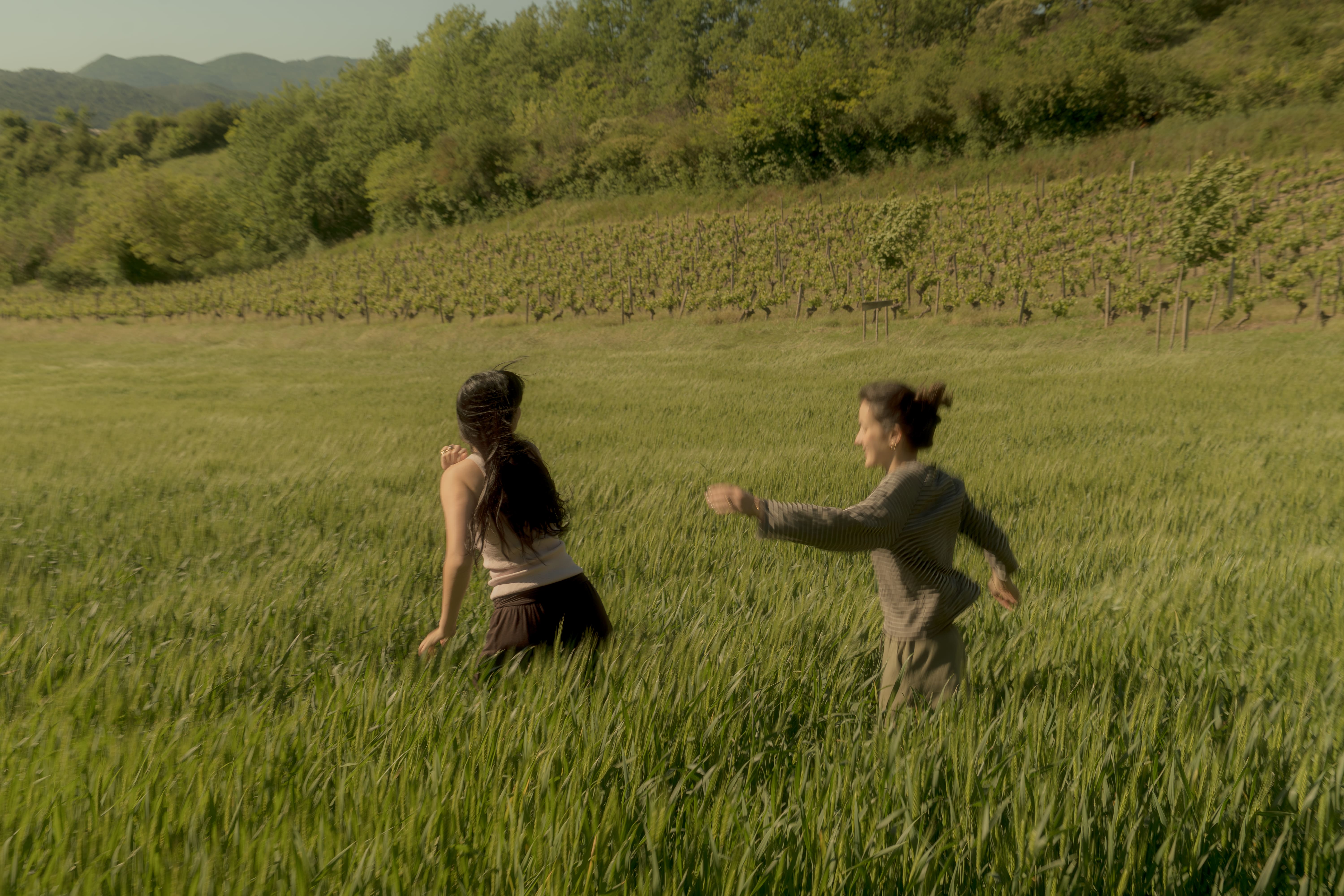 Two women in a grassy field exchanging small flowers on a bright, cloudy day.