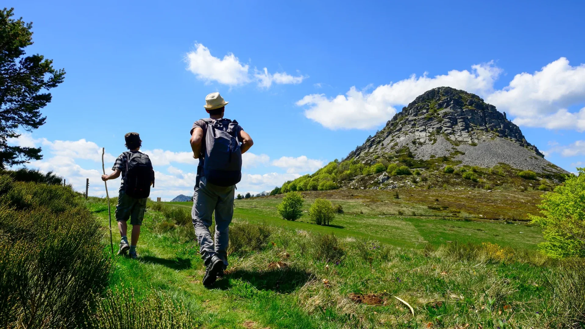 Deux randonneurs avec sac à dos marchent sur un sentier herbeux vers une colline rocheuse sous un ciel bleu.