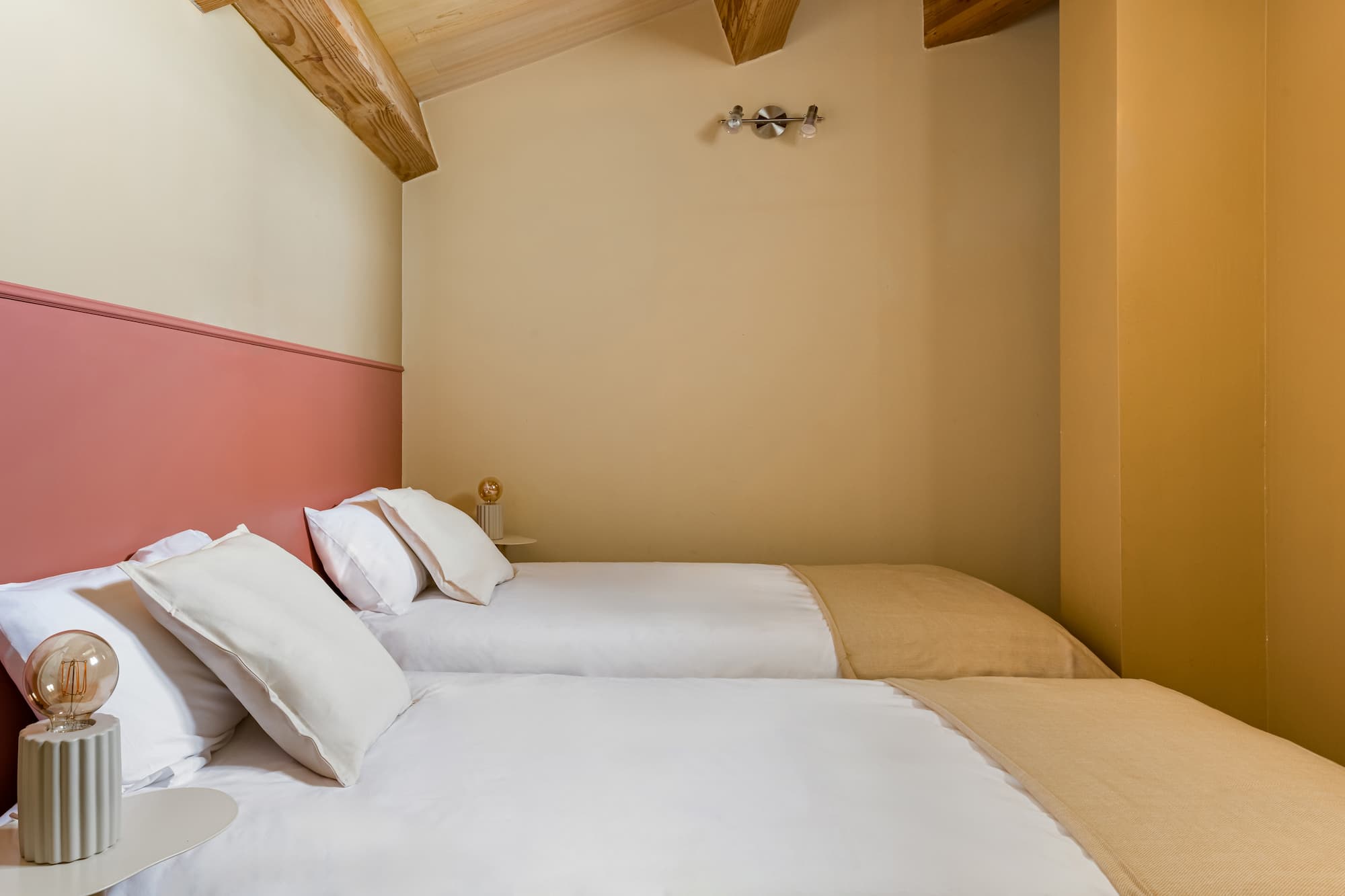 Chambre avec deux lits simples blancs, coussins, murs beige et poutres en bois au plafond.