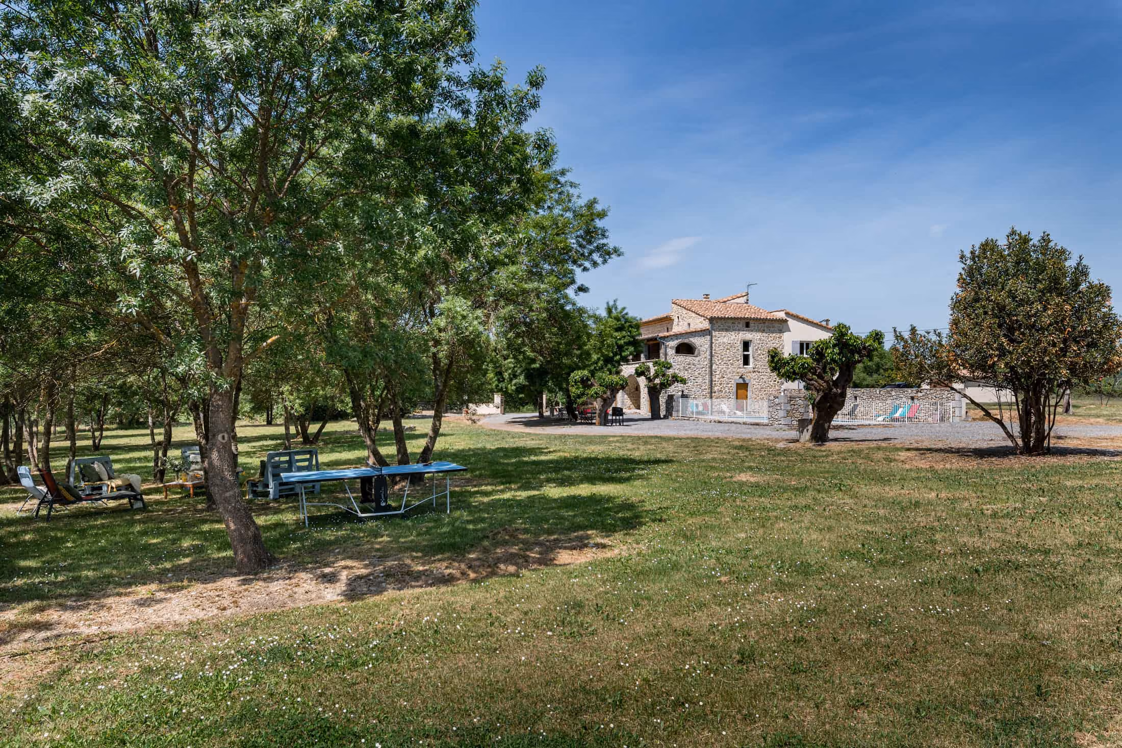 Grande maison en pierre avec terrain herbeux, arbres, table de ping-pong et chaises sous le soleil.