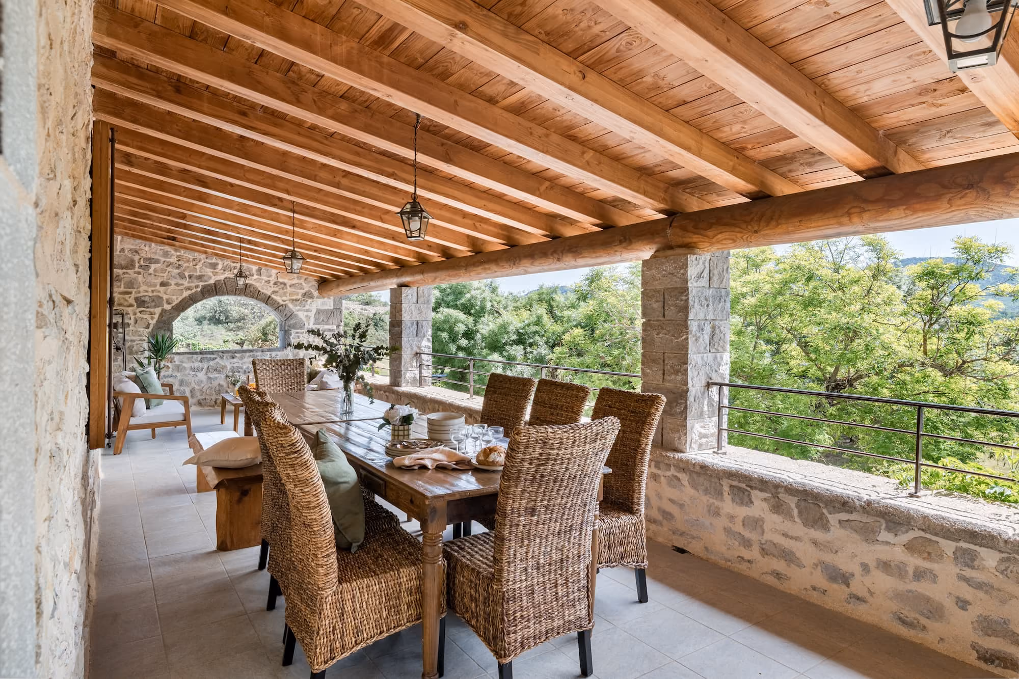 Terrasse avec table en bois, chaises en osier, plafond à poutres en bois et vue sur des arbres verts.
