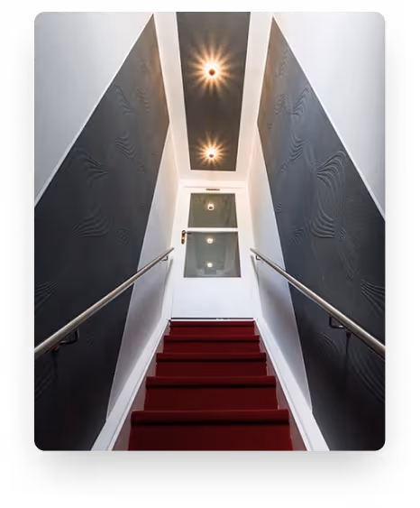 Treppenhaus mit roten Teppichstufen, weißen Wänden, schwarzen Wandpaneelen und Deckenlampen.