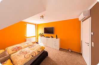 Gemütliches Schlafzimmer mit oranger Wand, Bett, TV auf Sideboard, Teppichboden und Klimaanlage.