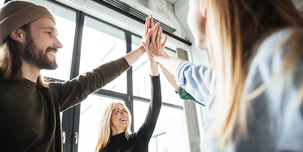 Three people smiling and giving a high five near large windows in a bright room.