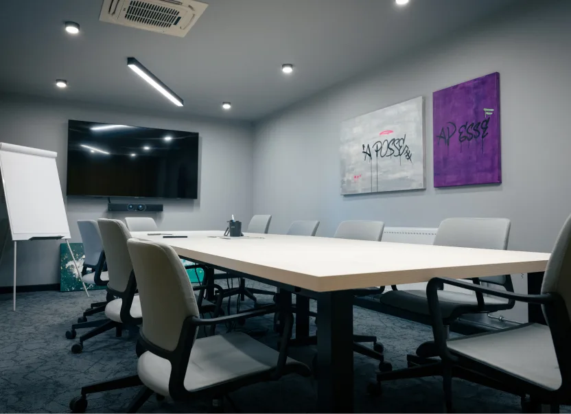 Modern conference room with a white table, gray chairs, TV screen, flip chart, and abstract wall art.