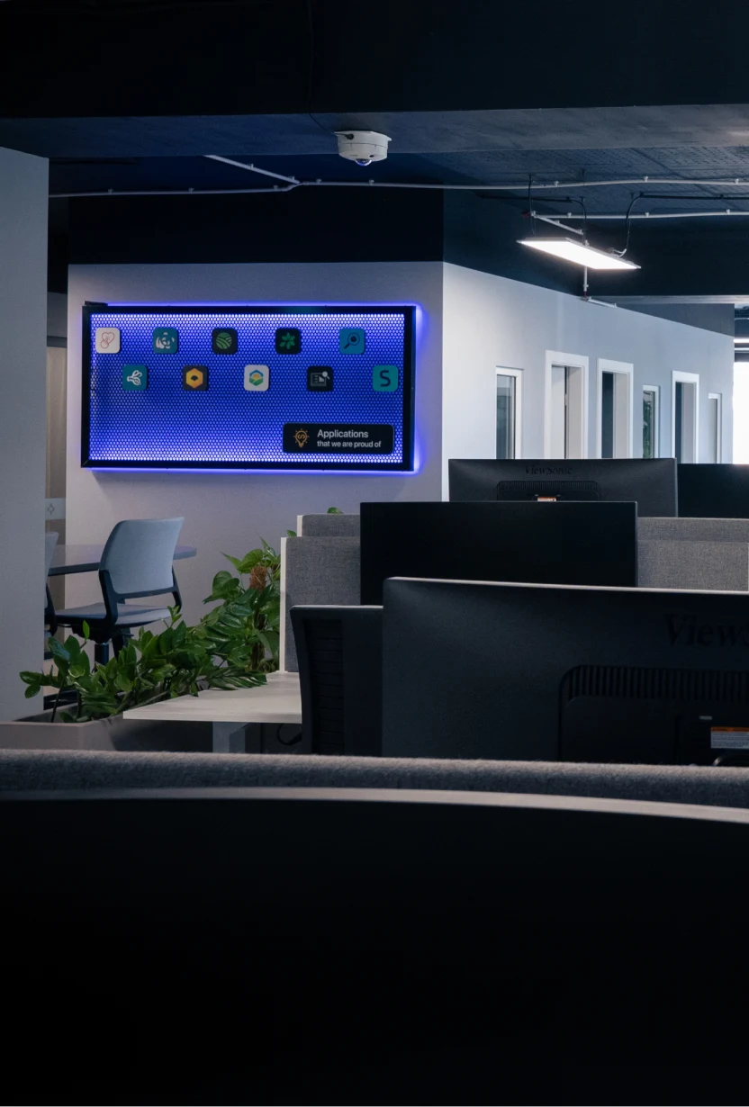 Modern office space with computer monitors, gray cubicles, chairs, plants, and a lit screen on a wall.