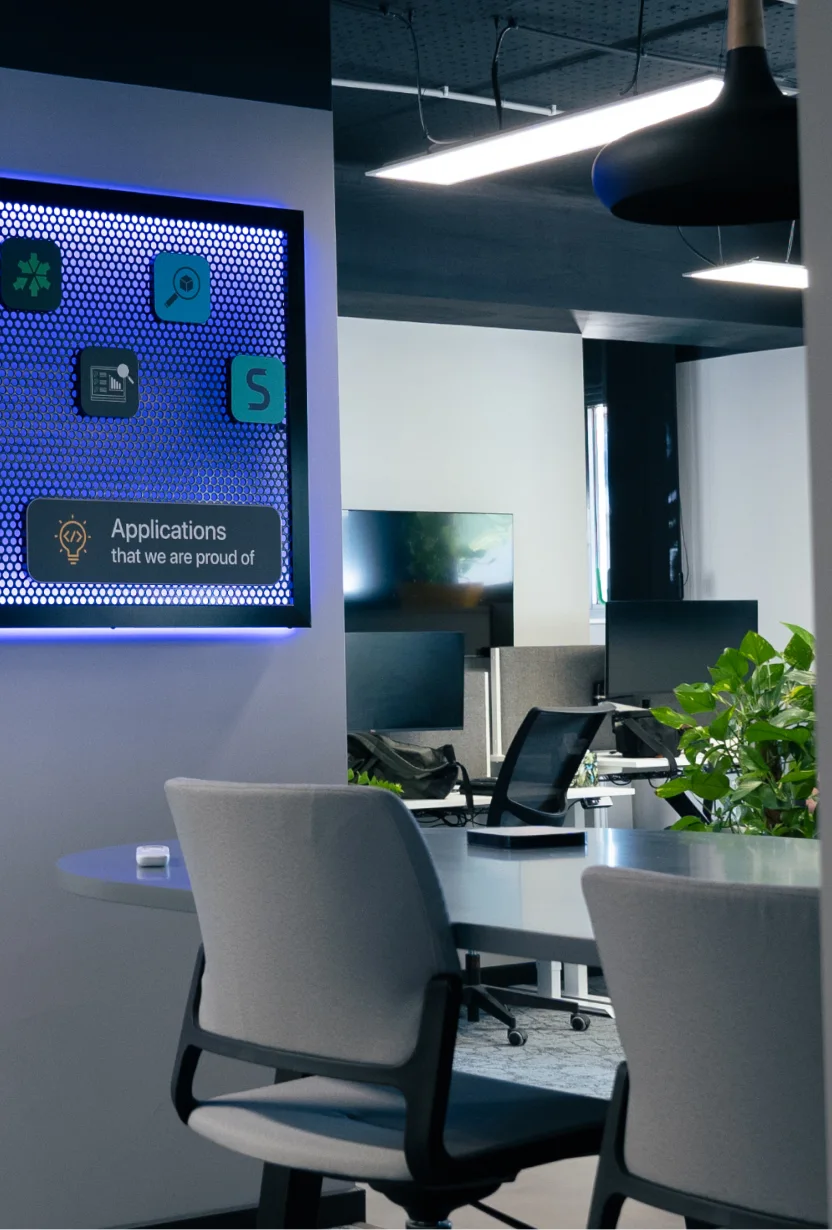 Modern office space with chairs, desks, monitors, plants, and a blue-lit screen saying Applications we are proud of.