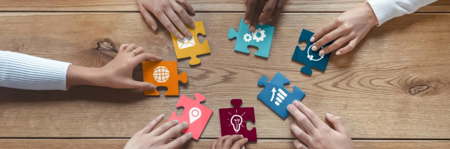 Multiple hands placing colorful puzzle pieces with icons representing communication and ideas on a wood table.