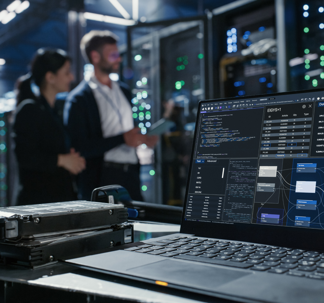A laptop displaying programming code and system data sits on a cart in a server room, with two people standing and talking in the background.