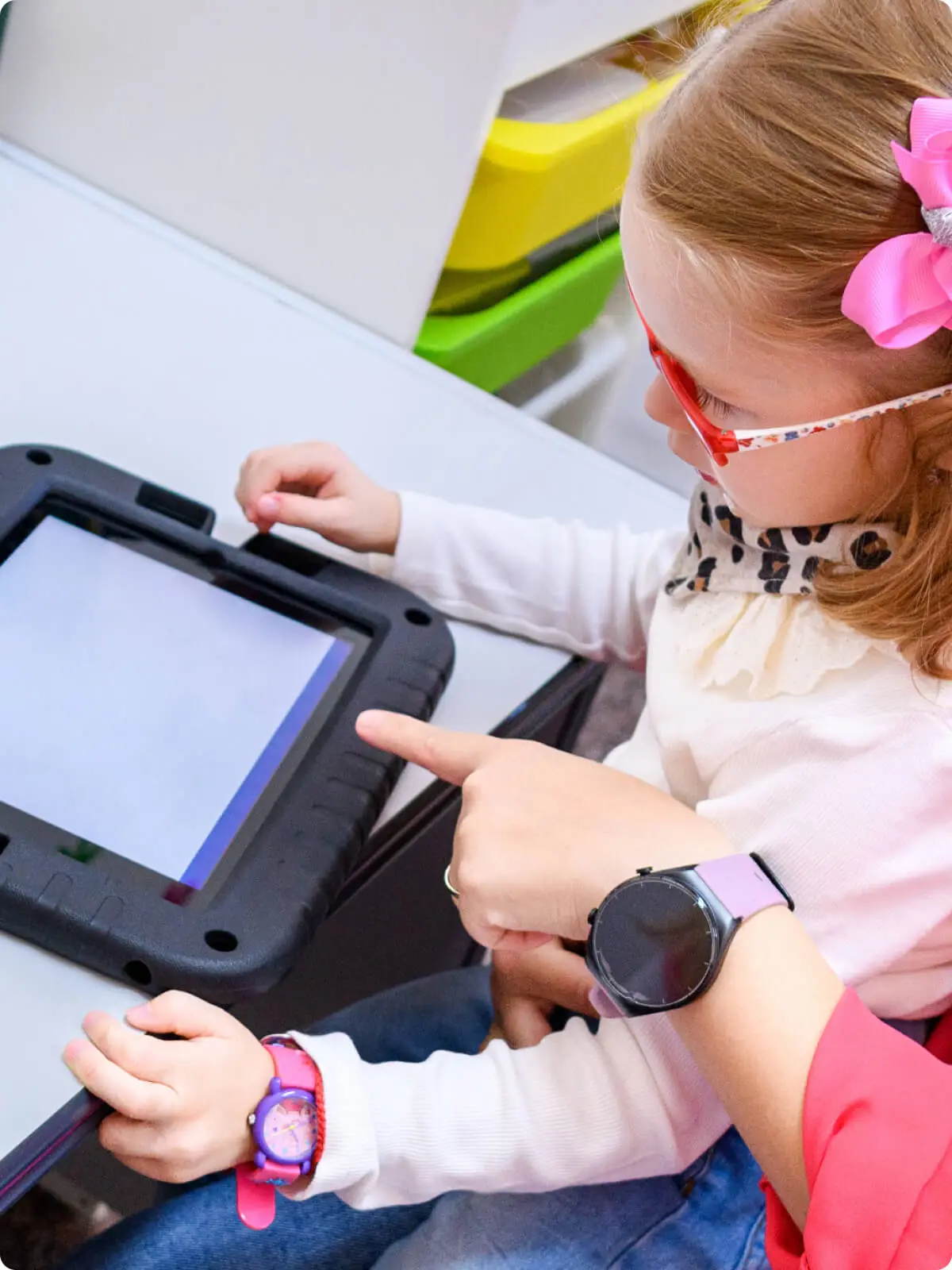 Young girl wearing glasses pointing at a rugged children's tablet in a classroom