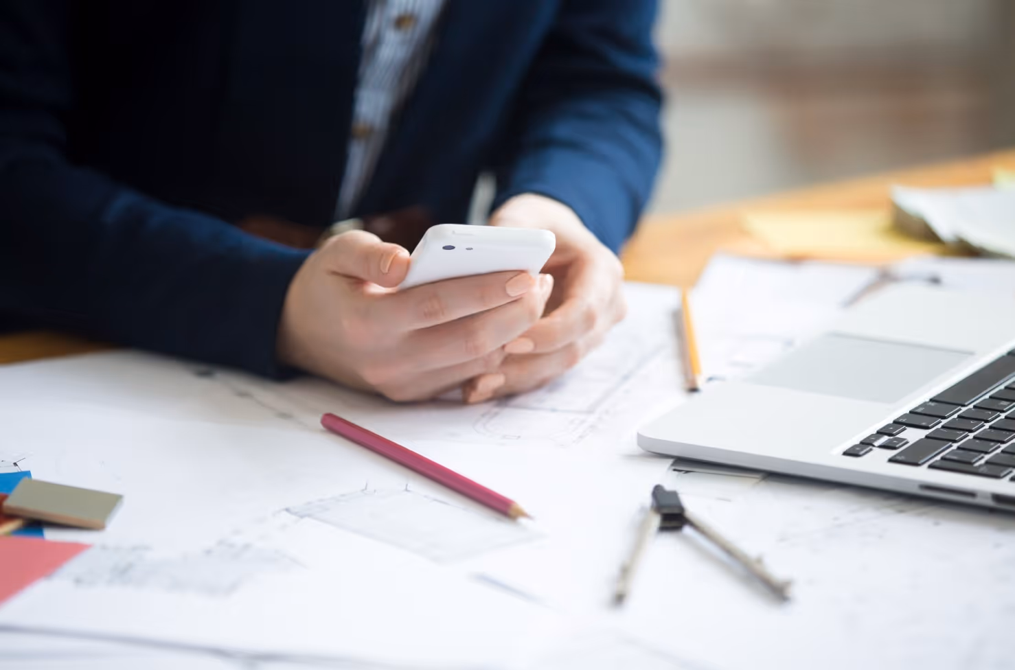 Személy fehér telefont tart kezében egy asztalon műszaki rajzokkal és laptopbal.