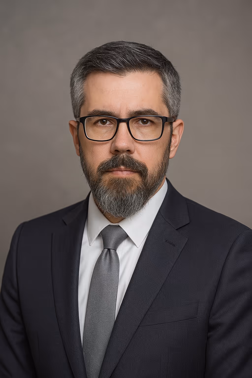 Komoly férfi szemüvegben sötét öltönyben, fehér inggel és szürke nyakkendővel szemben.