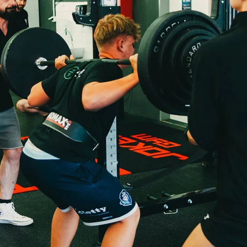Dřep s činkou v Joker's Gym, vedle stojí jistič