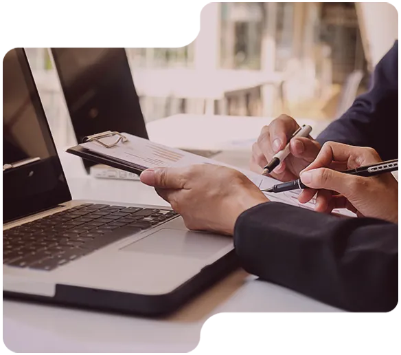 Zwei Personen überprüfen Dokumente mit Clip und Stiften vor einem Laptop.