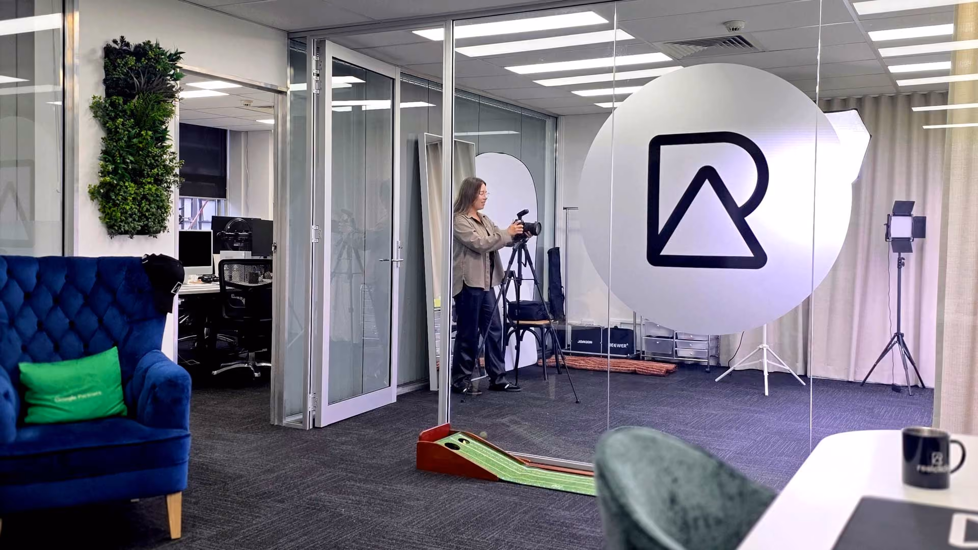 RealClicks team member adjusting camera on tripod in modern office studio with large logo on glass wall.