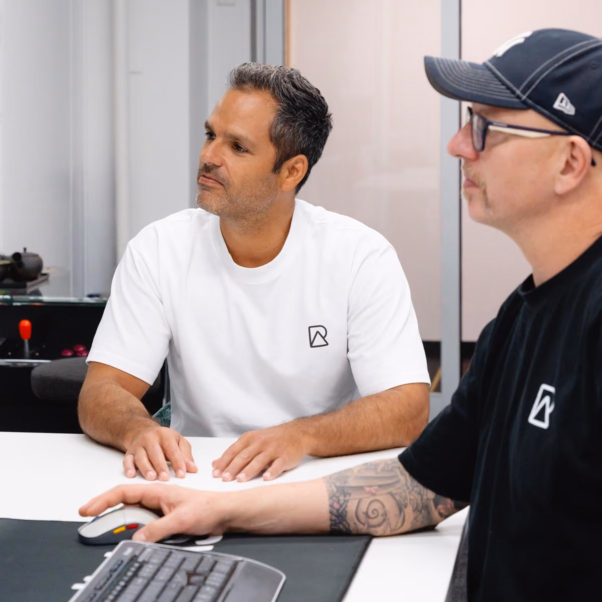 Two RealClicks team members at a desk, one using a mouse and keyboard, both wearing shirts with a logo.