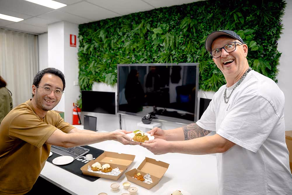 Two RealClicks team members smiling and exchanging a plate with food in an office with green wall and monitors.