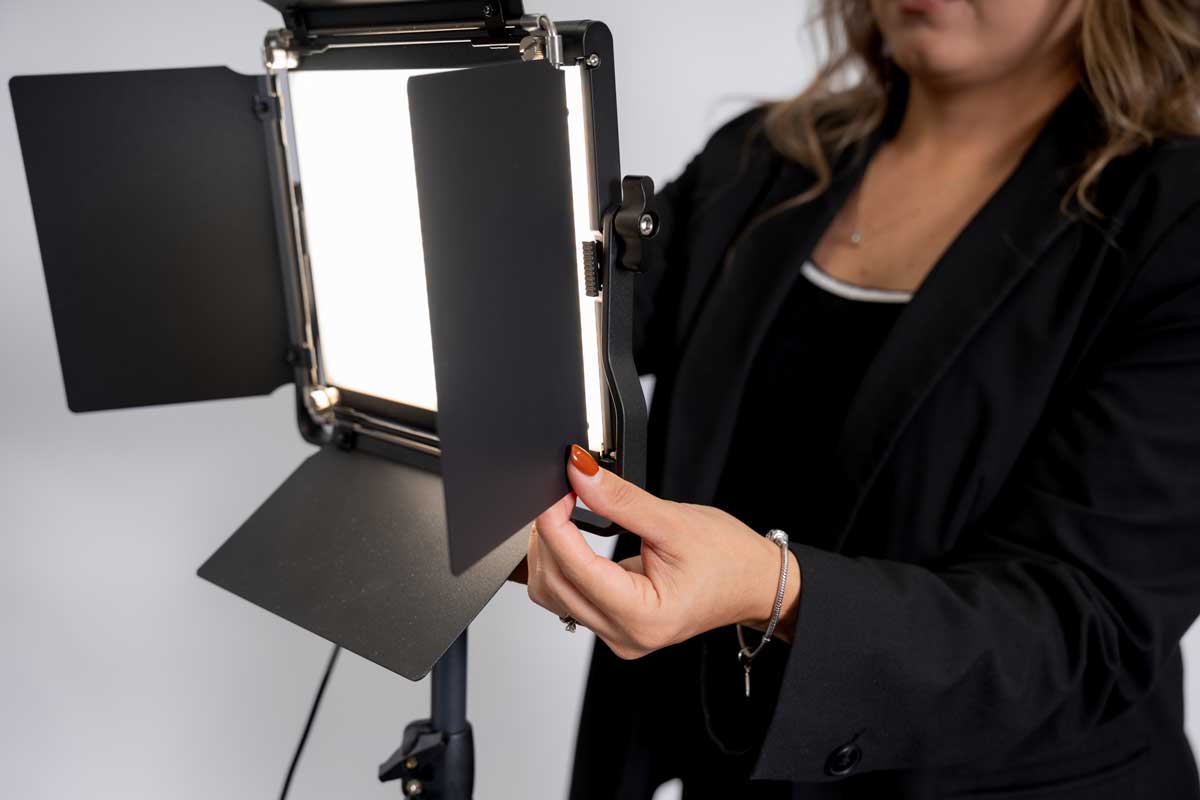A woman with manicured nails adjusts a studio light with open black barn doors on a tripod, wearing a black blazer and bracelet.