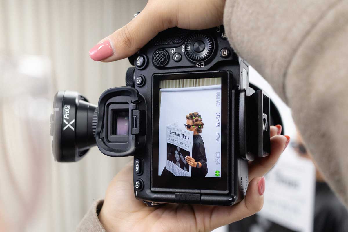 Hands holding a digital camera showing its screen, which displays a person with hair rollers wearing sunglasses and reading a newspaper titled 'Breaking News'.