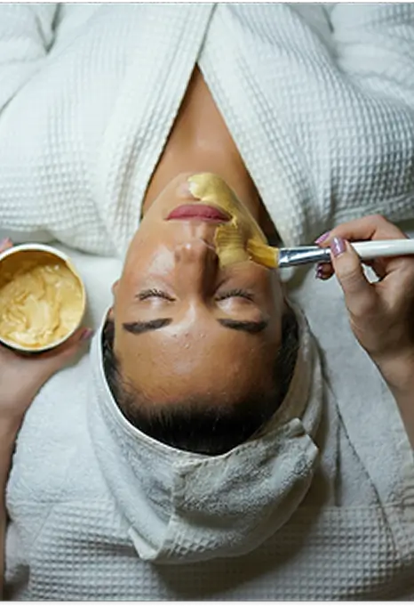 Mulher recebendo tratamento facial com máscara aplicada com pincel, olhos fechados, roupão branco.