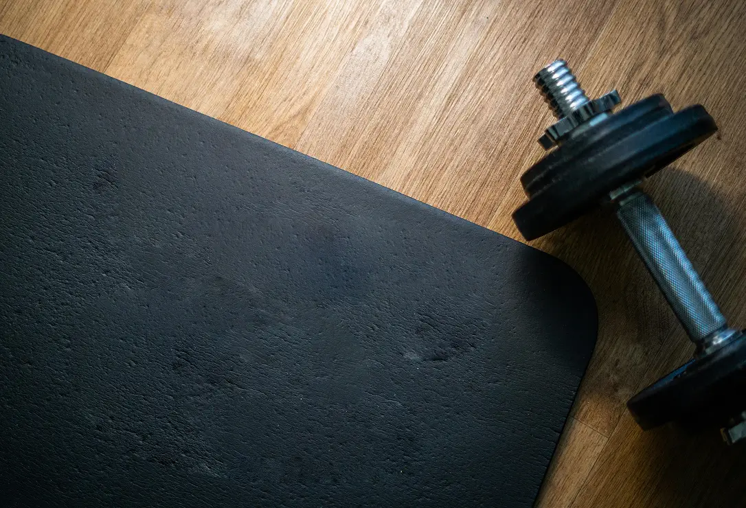 Um tapete de exercícios preto ao lado de um haltere com pesos em um piso de madeira.