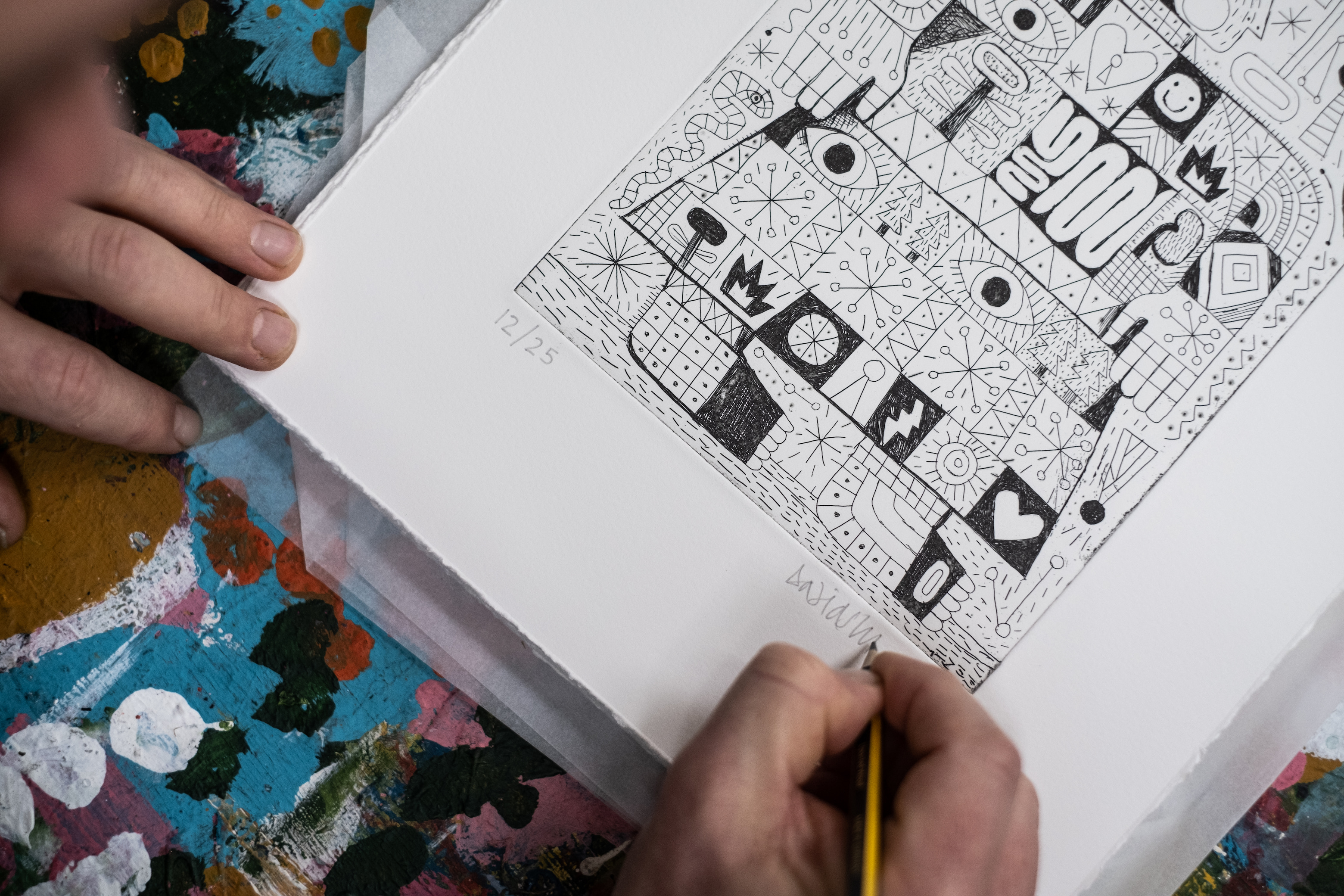David Shillinglaw pencil-signing a black-and-white Universal Humanoid etching (numbered 12/25) on the studio table, colourful paint-covered surface alongside.