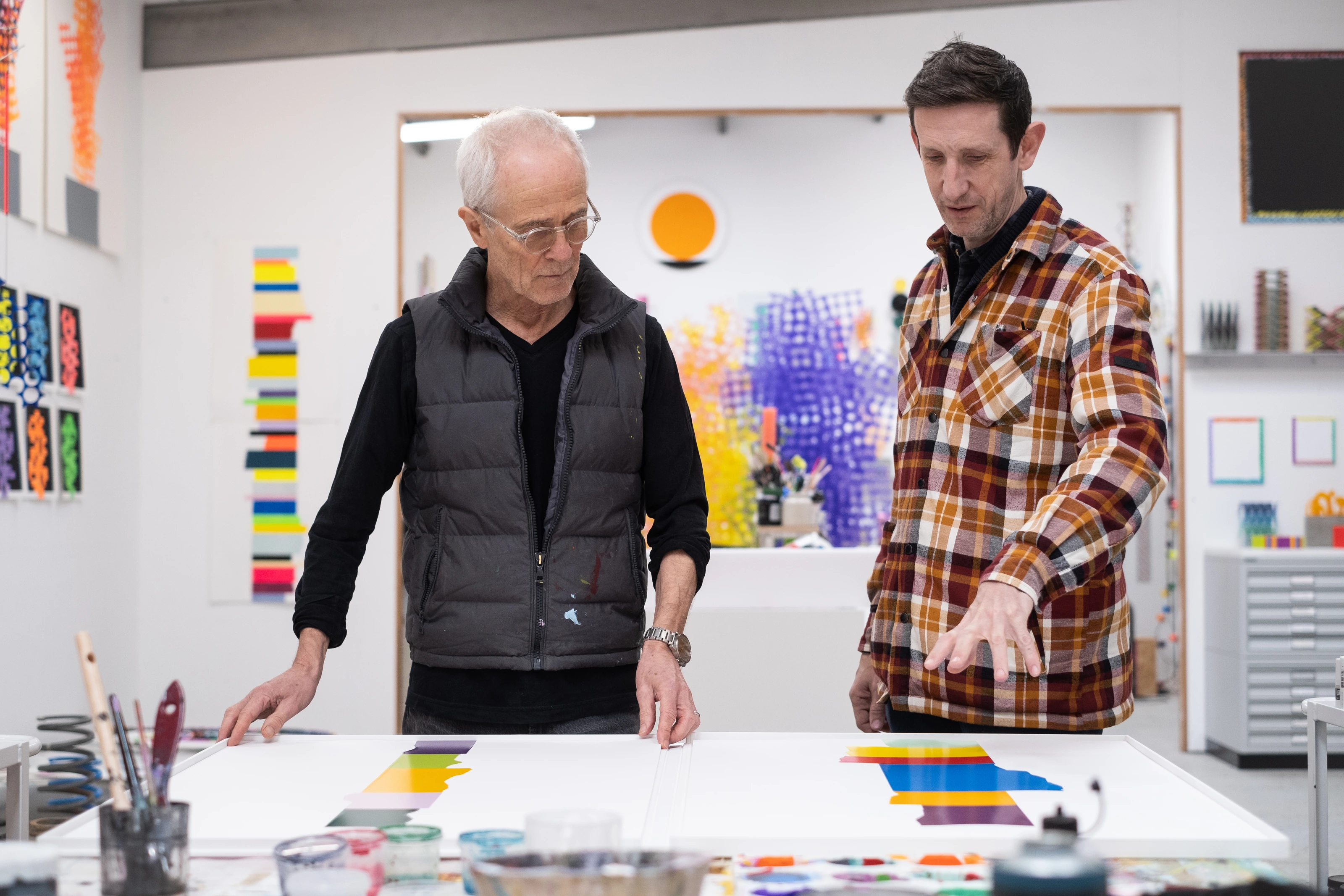 David Batchelor and master printer Peter Bennett studying two Chromophilia prints on a studio table, colourful artworks and materials filling the room behind them.