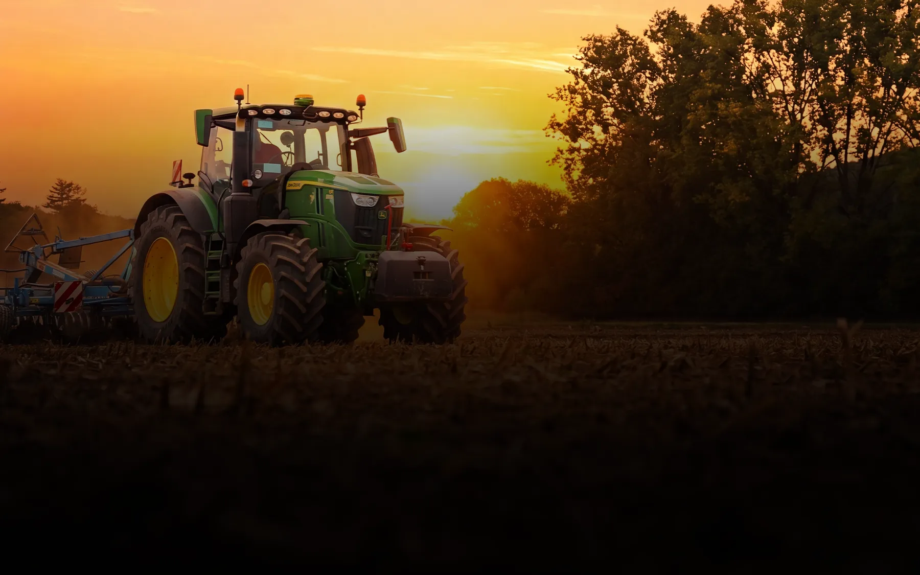 Grüner Traktor auf Feld bei Sonnenuntergang mit Bäumen im Hintergrund.