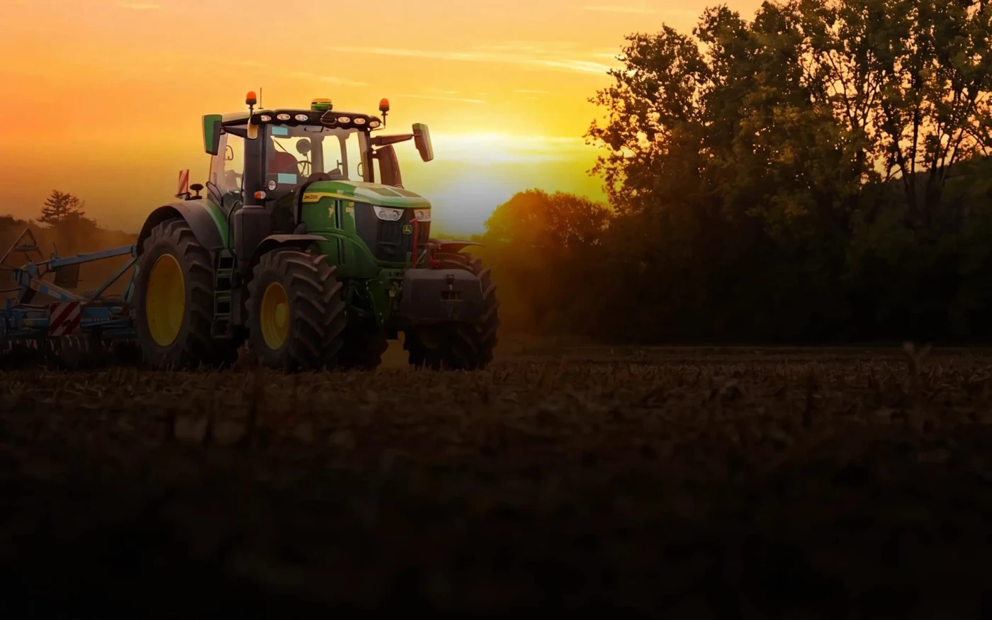Grüner Traktor auf Feld bei Sonnenuntergang mit Bäumen im Hintergrund.