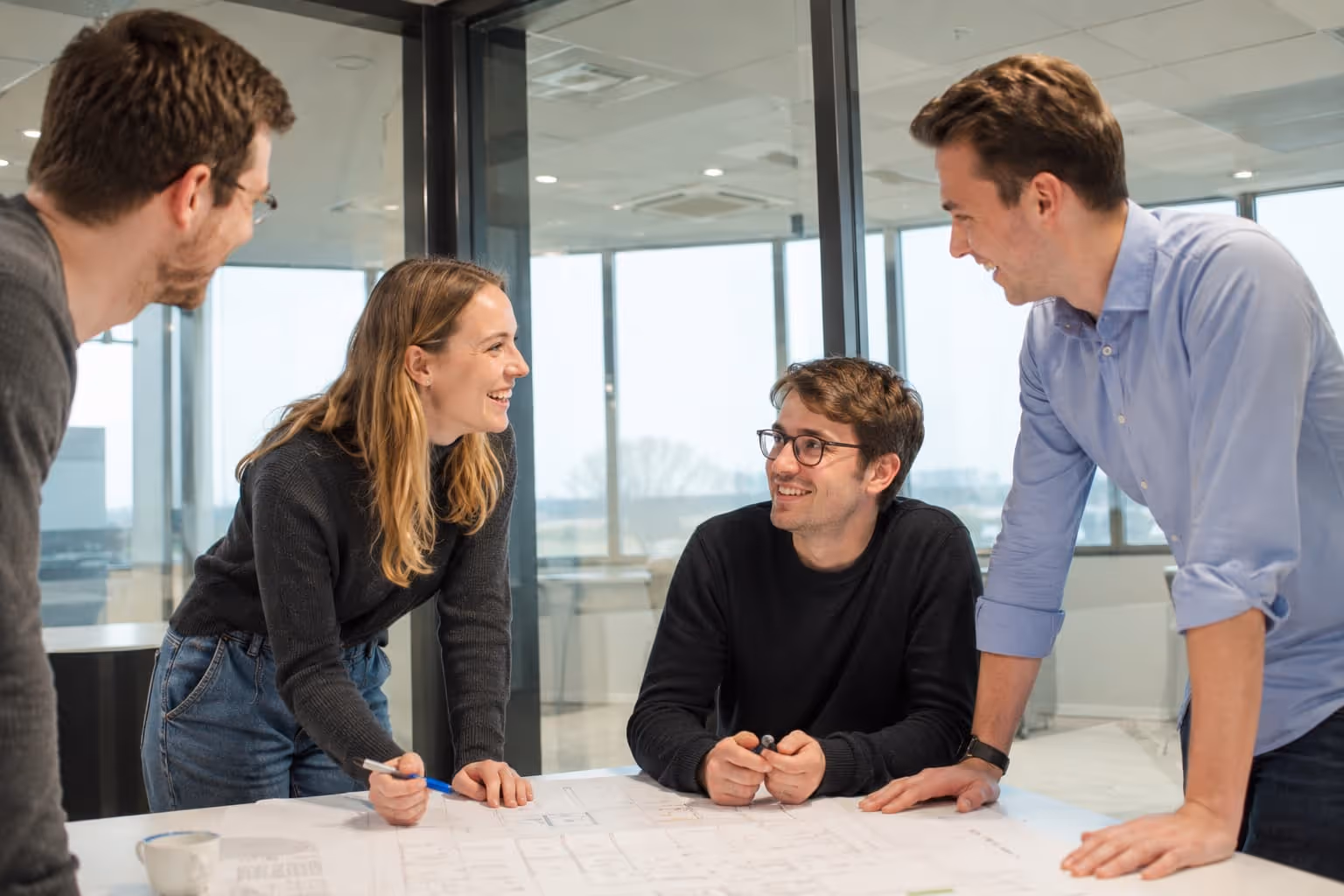 Trois collègues souriants discutant ensemble dans un bureau avec de grandes fenêtres.