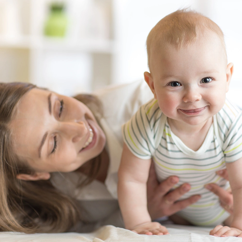 Lächelndes Baby in gestreiftem Strampler, das auf einem Bett krabbelt, während eine lächelnde Frau im Hintergrund das Baby stützt.