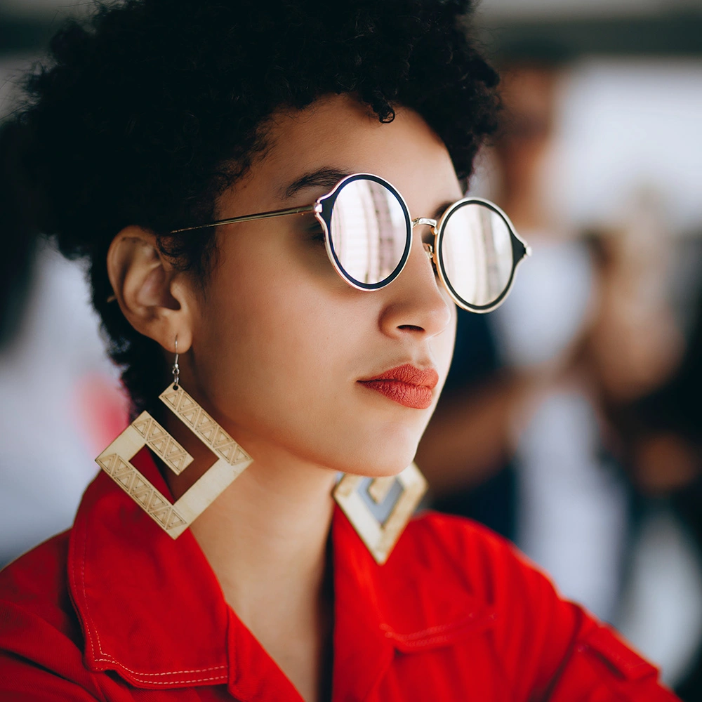 Junge Frau mit kurzen lockigen Haaren, rundgefasster Brille, roten Lippenstift, großen geometrischen Ohrringen und rotem Hemd.