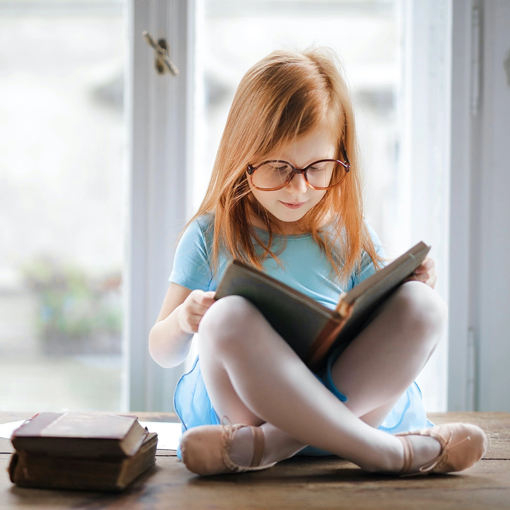 Ein rothaariges Mädchen mit Brille liest auf einem Holztisch sitzend ein Buch, neben ihr liegen zwei weitere Bücher.