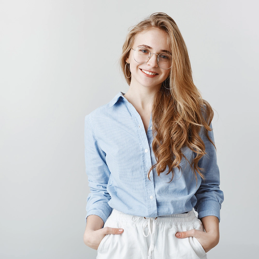 Junge Frau mit langen, welligen blonden Haaren, die helle Brille und blaues Hemd trägt und lächelt.