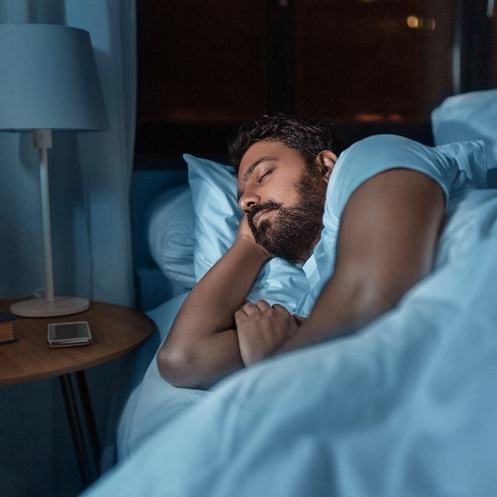 Mann schläft entspannend in einem Bett mit blauer Bettwäsche in einem dunklen Schlafzimmer.
