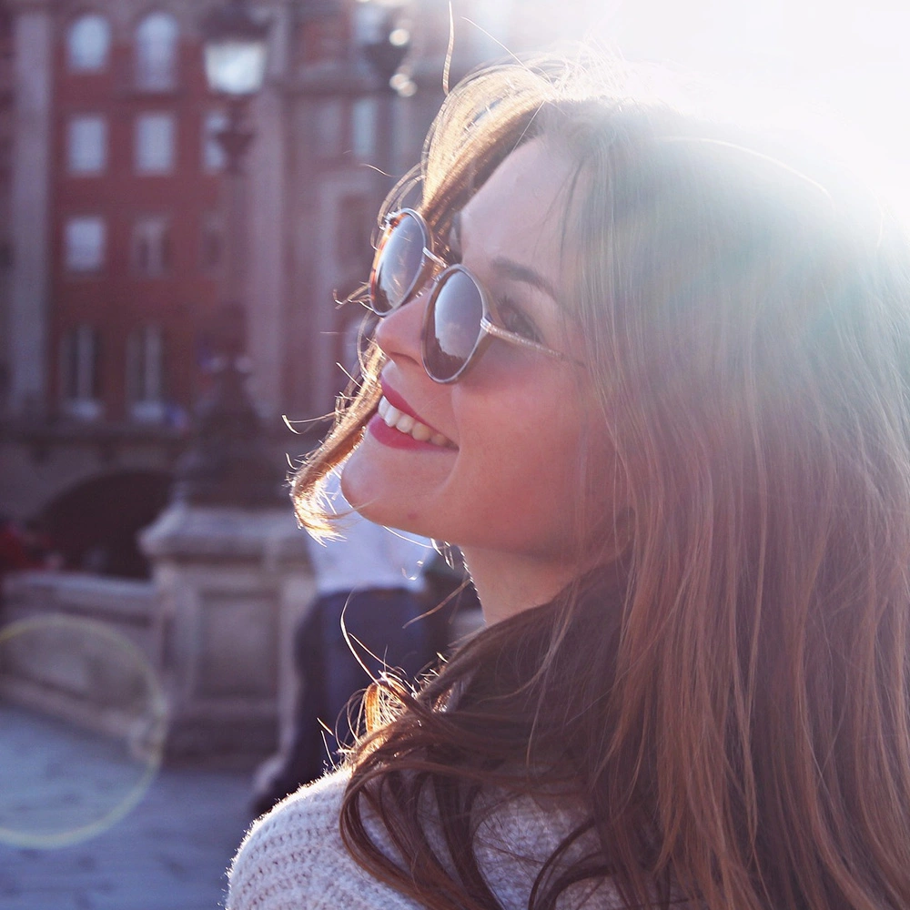 Frau mit langen braunen Haaren, die eine Sonnenbrille trägt und in der Sonne lächelt.