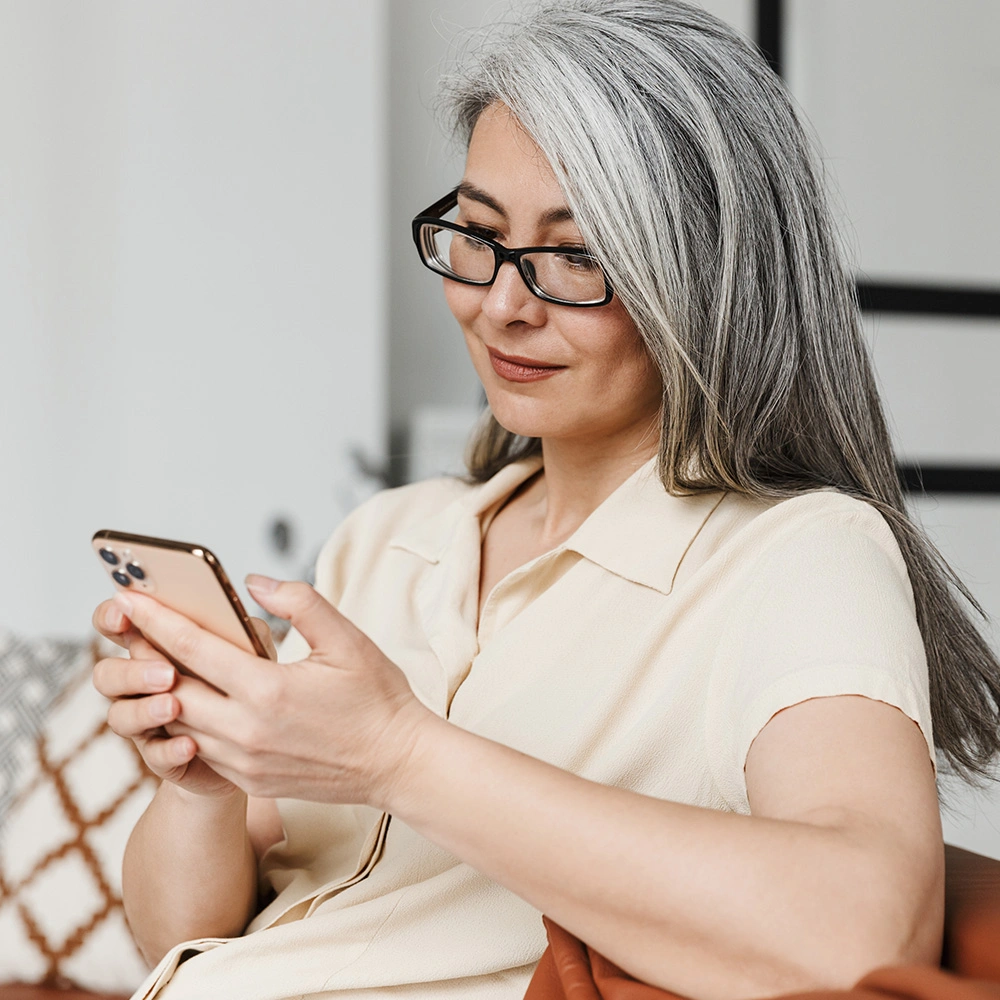 Frau mit grauem Haar und Brille sitzt auf einem Sofa und benutzt ein Smartphone.