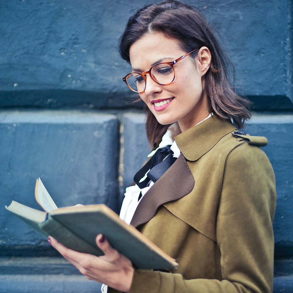 Frau mit brauner Brille und olivgrünem Mantel liest draußen ein Buch und lächelt.