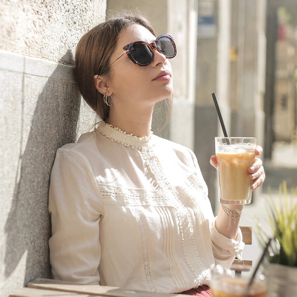 Frau mit Sonnenbrille lehnt an einer Wand und hält ein Glas mit Eiskaffee und Strohhalm.