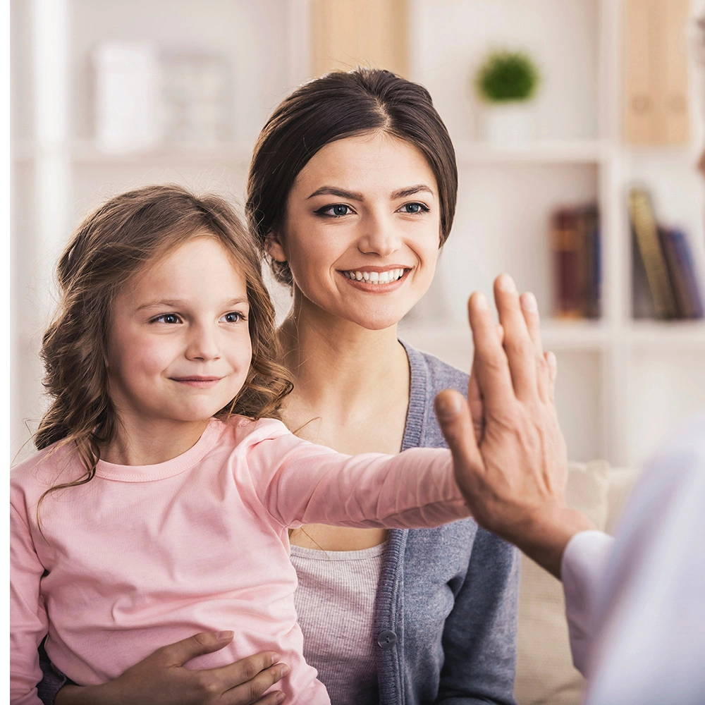 Mutter hält lächelnd ihre Tochter, die der Hand einer anderen Person zum High Five entgegenreckt.