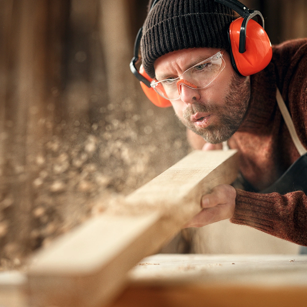 Mann trägt Gehörschutz und Schutzbrille und bläst Holzstaub von einem Holzbrett ab.