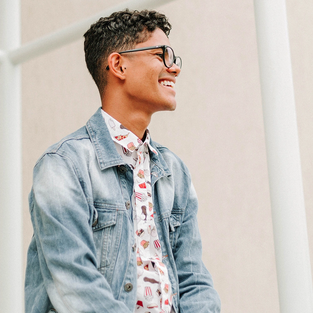 Junger Mann mit lockigem Haar und Brille, der lächelt und eine Jeansjacke über einem weißen Hemd mit Fast-Food-Motiven trägt.